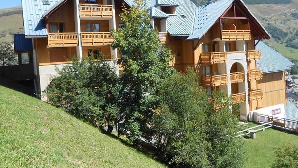 A multi-story wooden chalet with balconies, set against a mountainous backdrop.