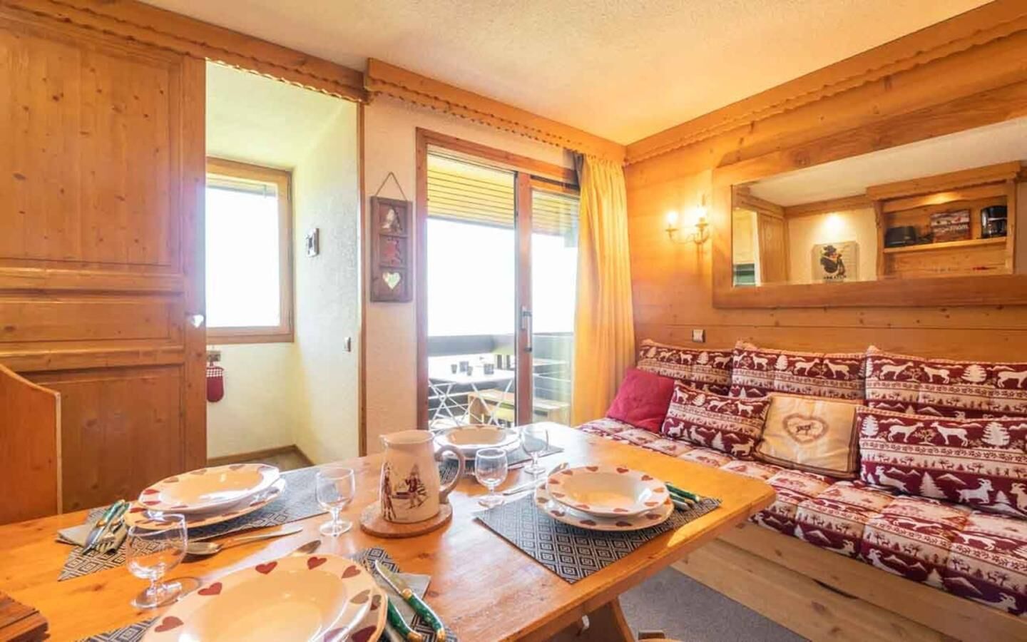 A cozy wooden interior with a dining table set for two, a sofa with patterned cushions, and a balcony with a view.