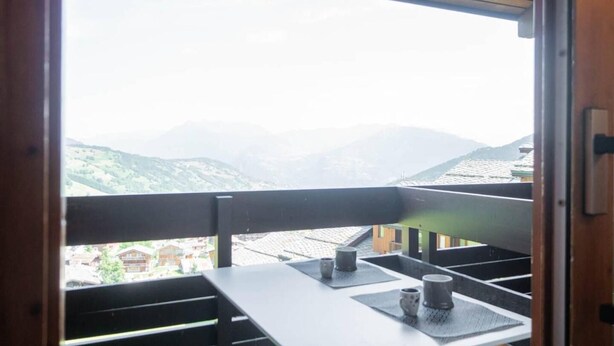 Een balkon met uitzicht op bergen, een tafel met twee kopjes, en een houten leuning.