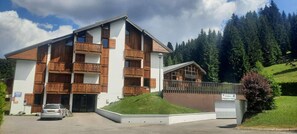 Un edificio de varios pisos con balcones de madera, un estacionamiento y montañas al fondo.