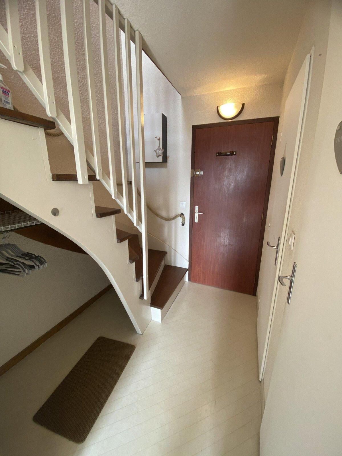 A narrow hallway with a wooden door, a small light fixture above it, and a staircase with white railings and wooden steps.