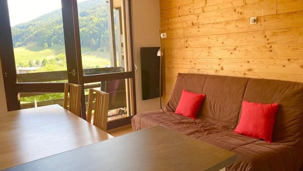 A cozy room with a wooden wall, a brown sofa with red cushions, a wooden table, and a large window offering a view of green hills. - 5th floor, balcony, tv, 43m², Villard de Lans (Villard-de-Lans)