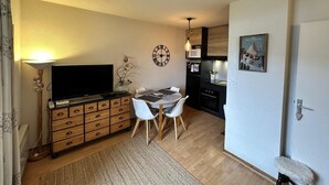 Un espacio habitable compacto con un televisor de pantalla plana sobre un mueble de madera, una mesa de comedor con sillas, una zona de cocina con microondas y una puerta con pomo en forma de corazón.