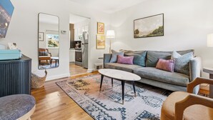 Ein Wohnzimmer mit einer Couch, einem Couchtisch und einem Teppich. Im Hintergrund ist eine Küche erkennbar.