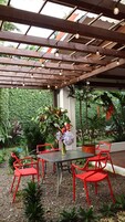 Un espacio de estar al aire libre cubierto con una mesa y sillas rojas, rodeado de abundante follaje y una pérgola de metal con luces colgantes.