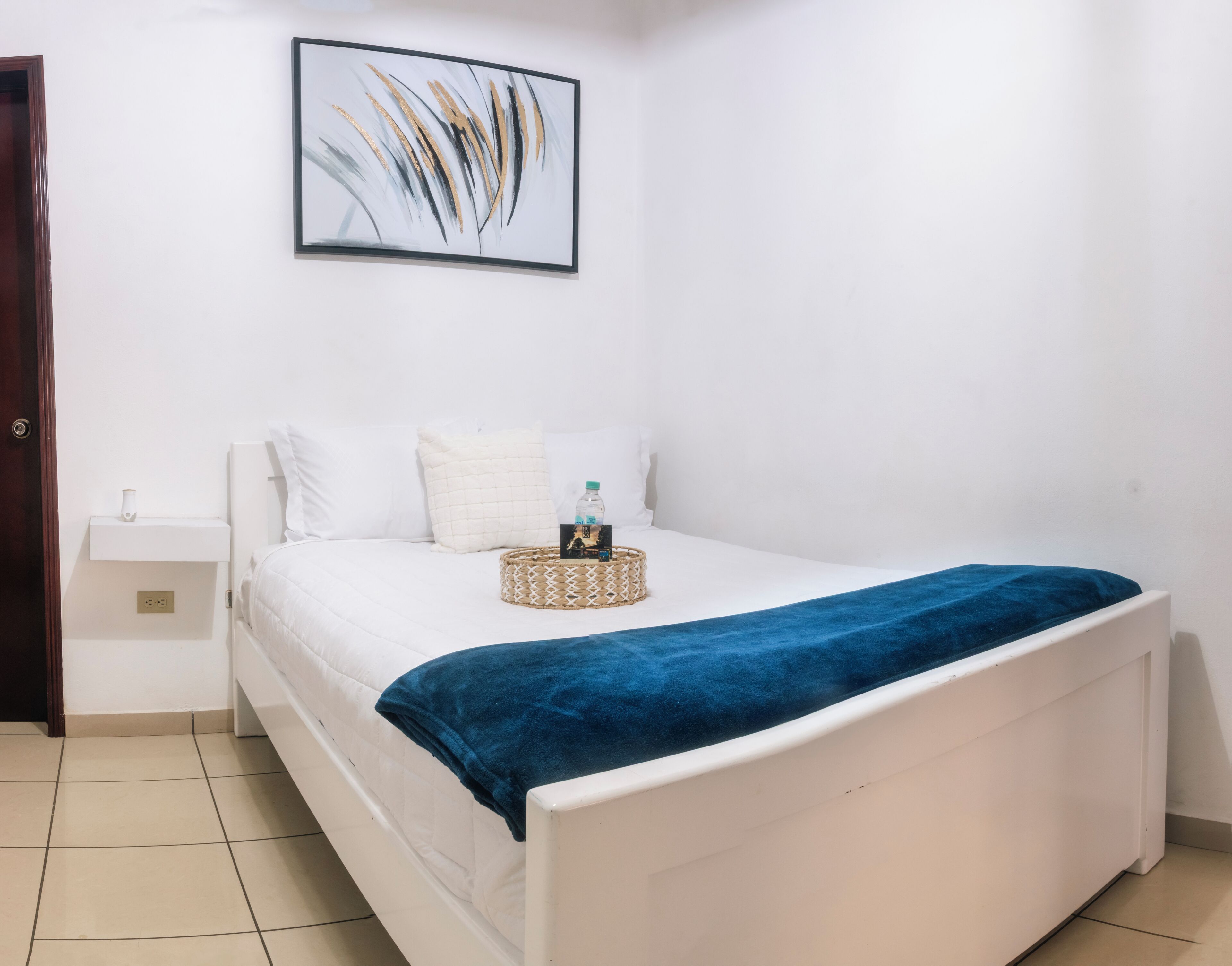 A bedroom with a bed, white walls, a framed abstract painting, a bedside table, and a blue blanket.