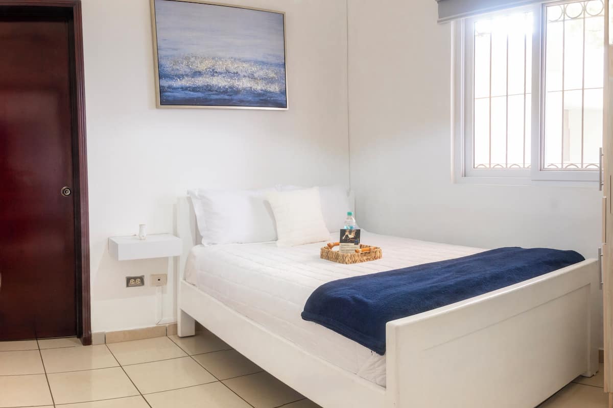 A bedroom with a white bed, a blue blanket, a bedside table with a bottle, and a painting on the wall.