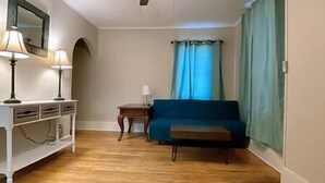 Een kamer met een houten vloer, een blauwe bank, een kleine tafel met een lamp, een plafondventilator en een raam met gordijnen.