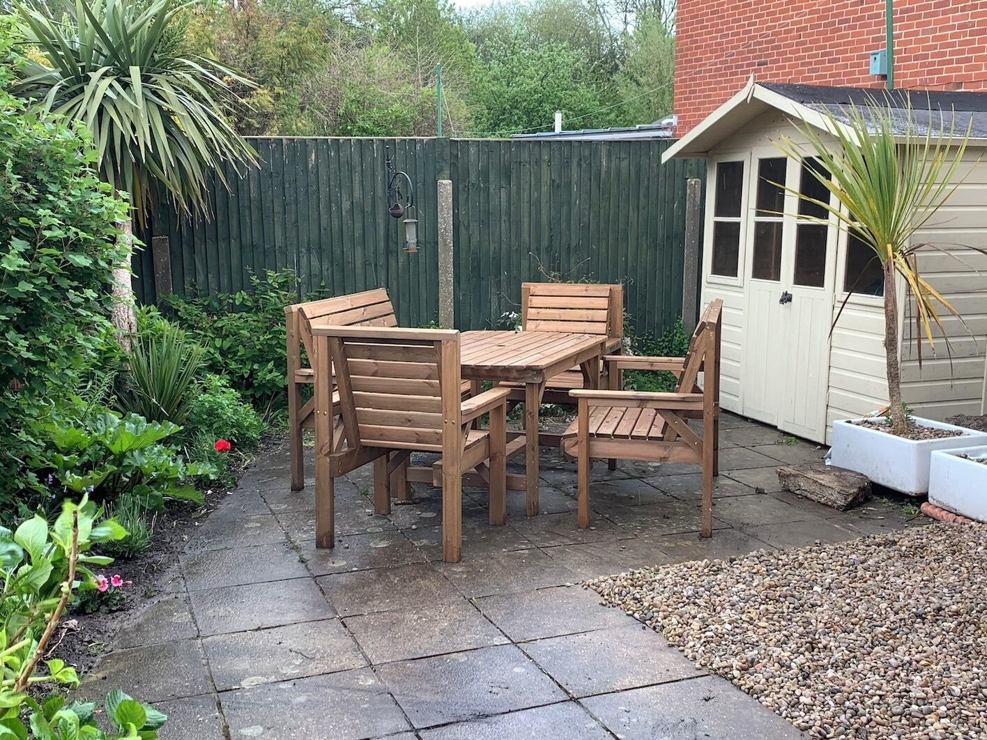 Eine Terrasse mit Holztisch und -stühlen, umgeben von Pflanzen und einem grünen Zaun.