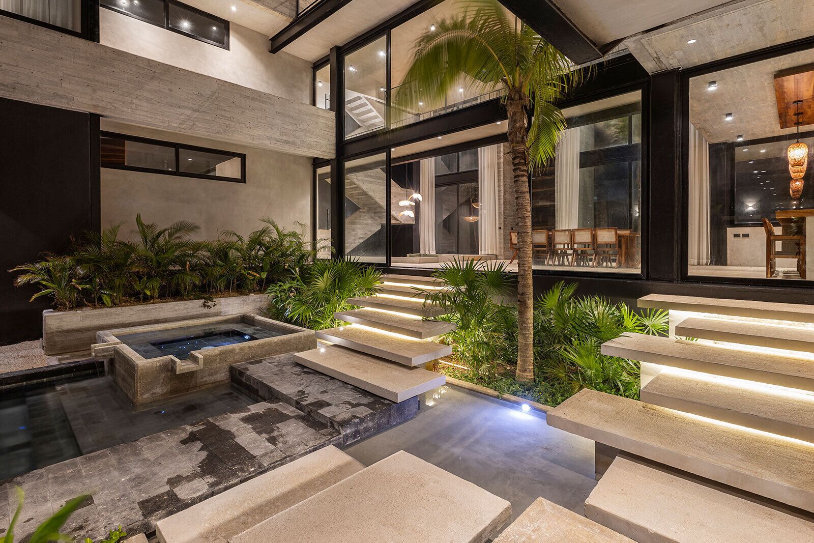 A modern indoor pool area with a staircase, palm trees, and a seating area.
