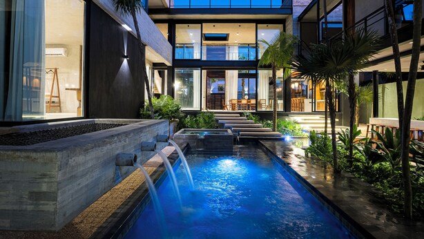 A modern outdoor pool area with a waterfall, surrounded by lush greenery and a contemporary building with large windows.