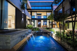 A modern outdoor pool area with a waterfall, surrounded by lush greenery and a contemporary building with large windows.
