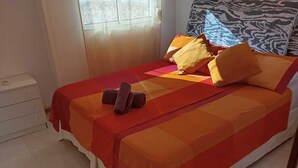 Een bed met een rood-oranje sprei, meerdere kussens en een paar opgevouwen handdoeken aan de zijkant.