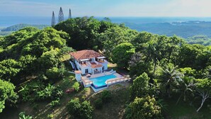 Ein großes, zweistöckiges Haus mit Swimmingpool, umgeben von üppiger Vegetation und Blick auf den Ozean.