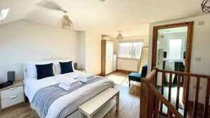 A modern bedroom with a large bed, bedside tables, a bench, and a view of the kitchen and balcony. - Hereford House – Sleeps 8! BS2 (Bristol)