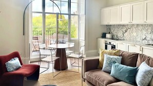 Una sala moderna con una mesa redonda y sillas, un sillón rojo, un sofá marrón con cojines, y una cocina con armarios blancos y un protector contra salpicaduras de mármol.