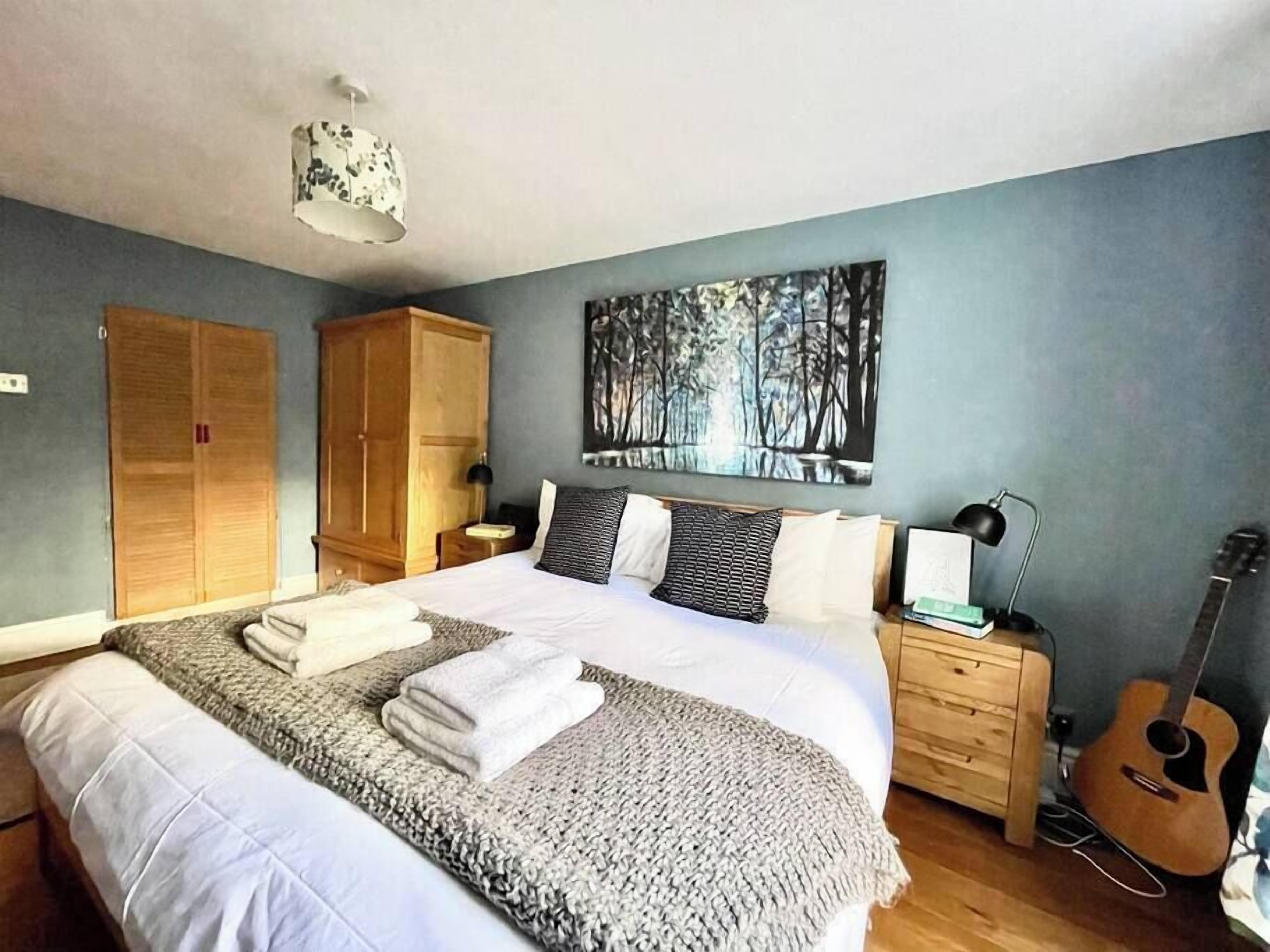 A bedroom with a large bed, a wardrobe, a guitar, and a painting on the wall.