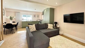 Un salon moderne avec un canapé d’angle gris, un coin repas avec une table et des chaises, une cuisine en arrière-plan et un téléviseur à écran plat fixé au mur.