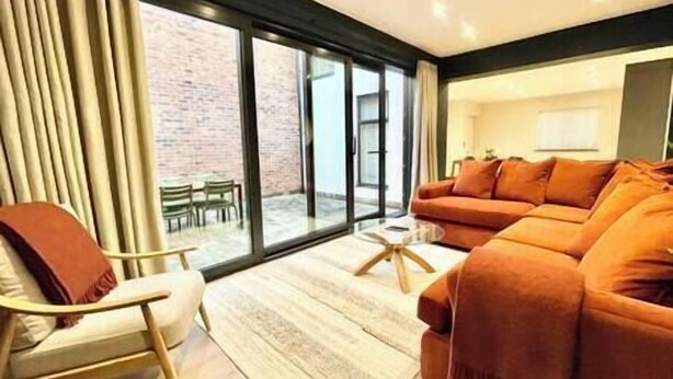 Un salon moderne avec un grand canapé, une table basse et un fauteuil. Une porte-fenêtre coulissante donne accès à un espace extérieur agrémenté d’un mur en briques et d’une table avec des chaises.