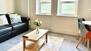 A living room with a black leather sofa, a wooden coffee table, a vase with flowers, and a dining area with patterned chairs. - New and Spacious Apartment - Sleeps 5 (Bristol)