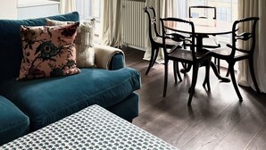 A living room with a blue sofa, a dining table with chairs, a chandelier, and large windows with curtains. - New King Street Suites - Apartment 1 (Bath)