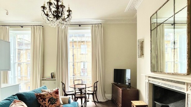 A living room with a blue sofa, a patterned ottoman, a chandelier, a fireplace, and a large mirror. - New King Street Suites - Apartment 1 (Bath)