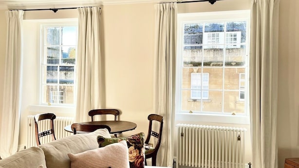 A cozy living room with a beige sofa, a patterned throw pillow, a white armchair, and a chandelier. There is a dining table with chairs and a view of the outside through large windows with curtains.