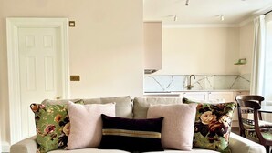 A living room with a beige sofa, floral cushions, and a white throw. A kitchen is visible in the background.