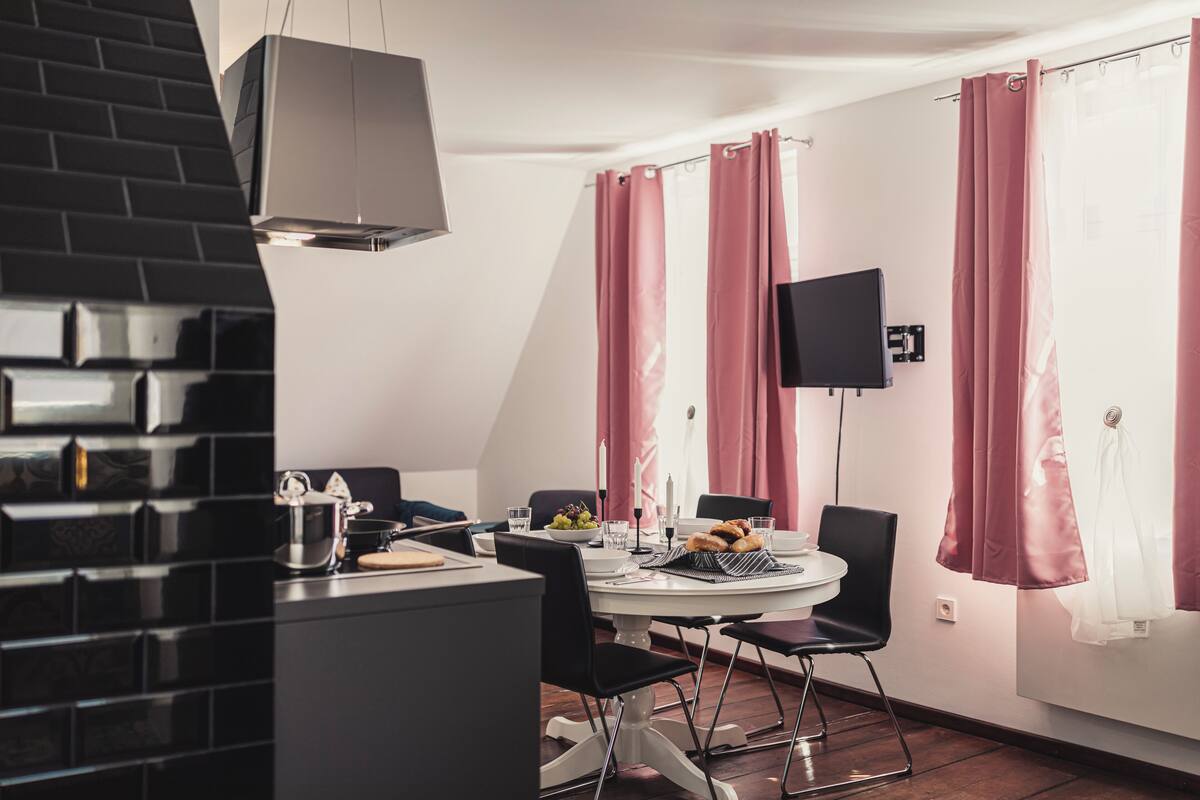 A modern dining area with a round table, black chairs, a TV mounted on the wall, and pink curtains.