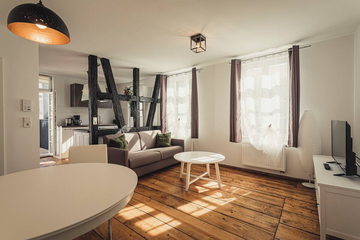 A modern living room with a wooden floor, a white sofa, a round dining table, and a kitchen in the background.