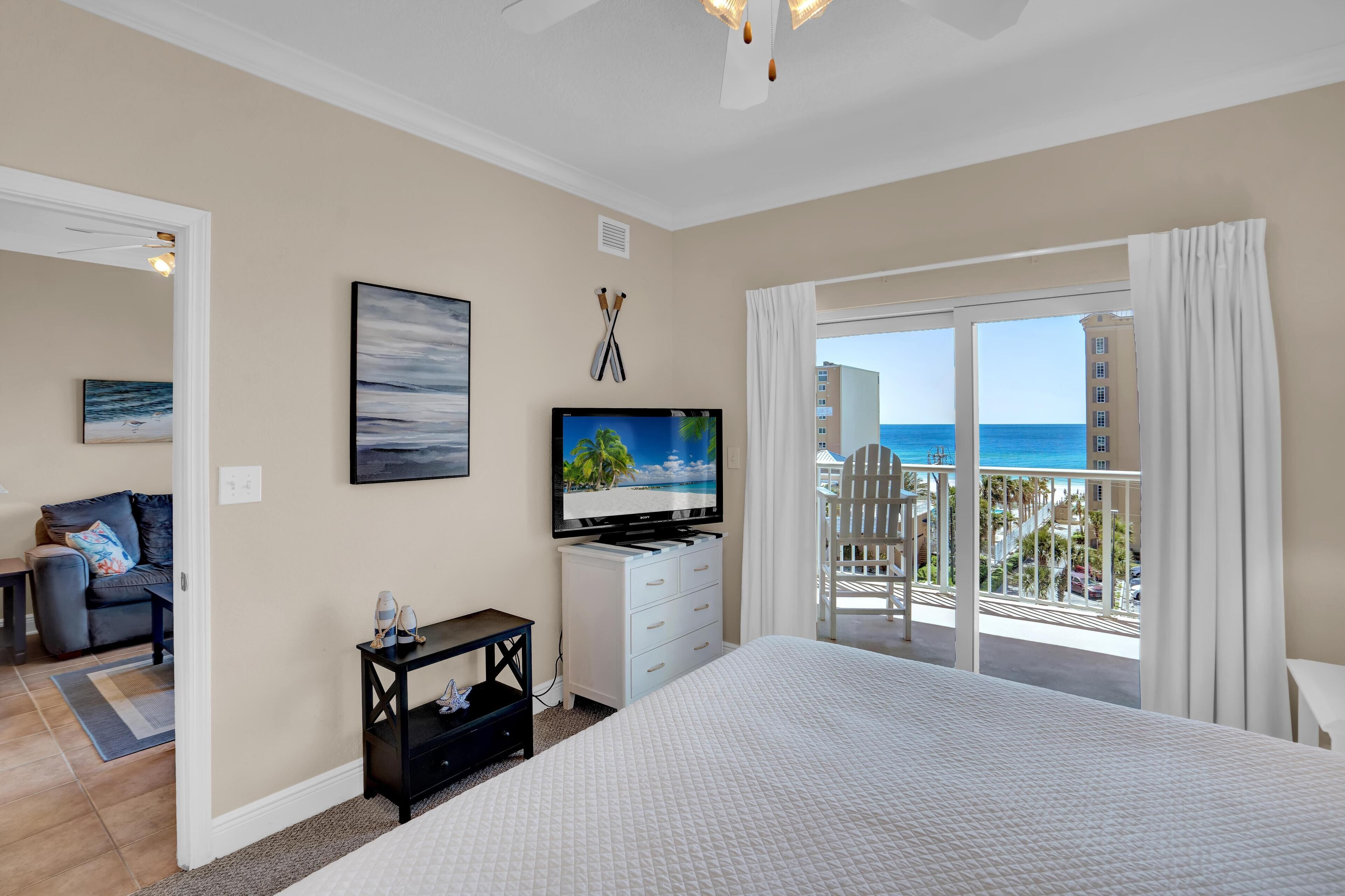 A bedroom with a large bed, a TV on a white dresser, a painting, and a balcony with a view of the ocean.
