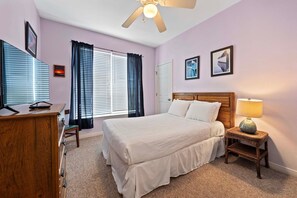 Ein Schlafzimmer mit einem Bett, einem Nachttisch mit Lampe, einem Schreibtisch mit Computer und einem Fenster mit Vorhängen.