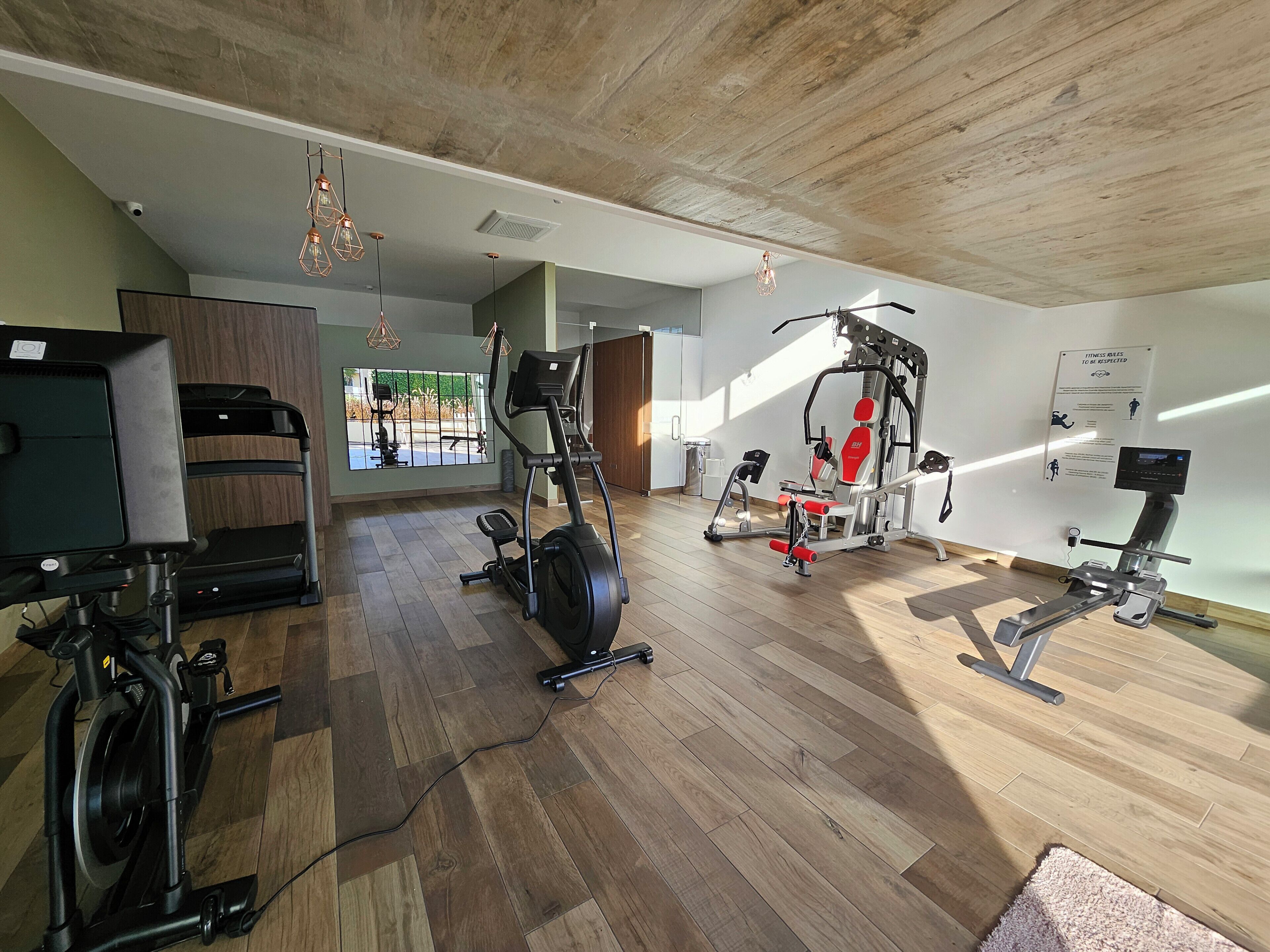 Ein geräumiger Fitnessraum mit verschiedenen Trainingsgeräten, Holzboden und großen Fenstern für natürliches Licht.