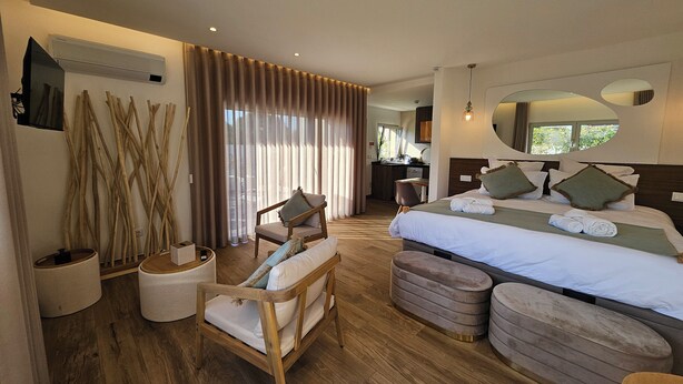 A modern hotel room with a large bed, a sitting area with a chair and ottoman, a wooden floor, and a view of the outside through the window. - Marinha Grande Apartamentos Rainbow N°9 (Marinha Grande)
