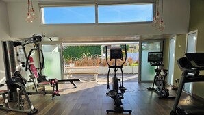 A gym with various exercise equipment, a bench, and a view of a garden through large windows. - Marinha Grande Apartamentos Rainbow N°9 (Marinha Grande)