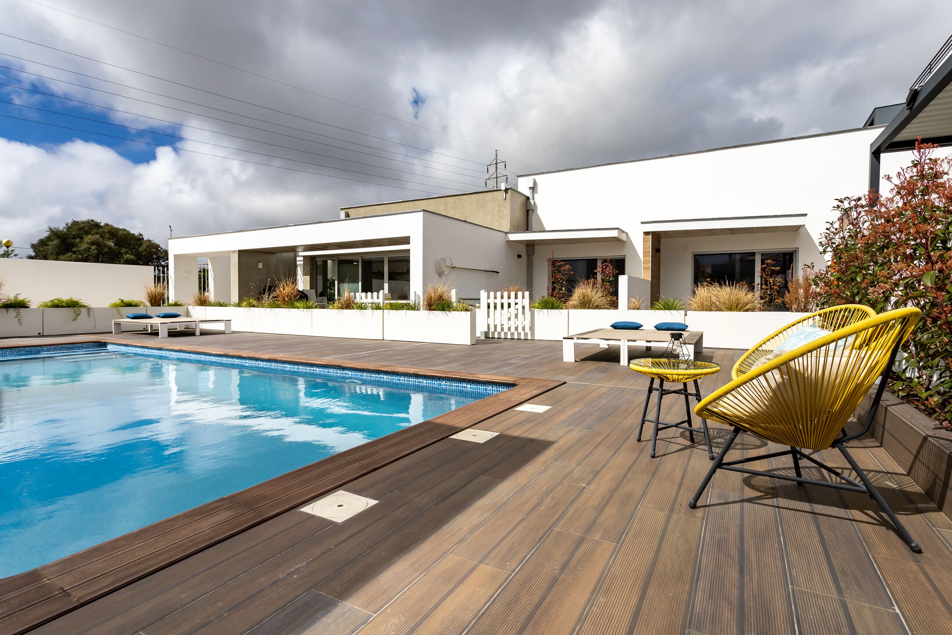 Uma área de piscina moderna ao ar livre, com um deck de madeira, um edifício branco e cadeiras amarelas.