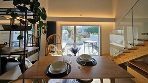 A modern dining area with a wooden table set for two, a wicker chair, and a glass staircase leading to a balcony. - Marinha Grande Apartamentos Duo N°7 (Marinha Grande)