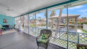A covered outdoor area with a wicker chair, a table with chairs, and a view of a pool and buildings in the background. - Fun In The Sun (Orange Beach)