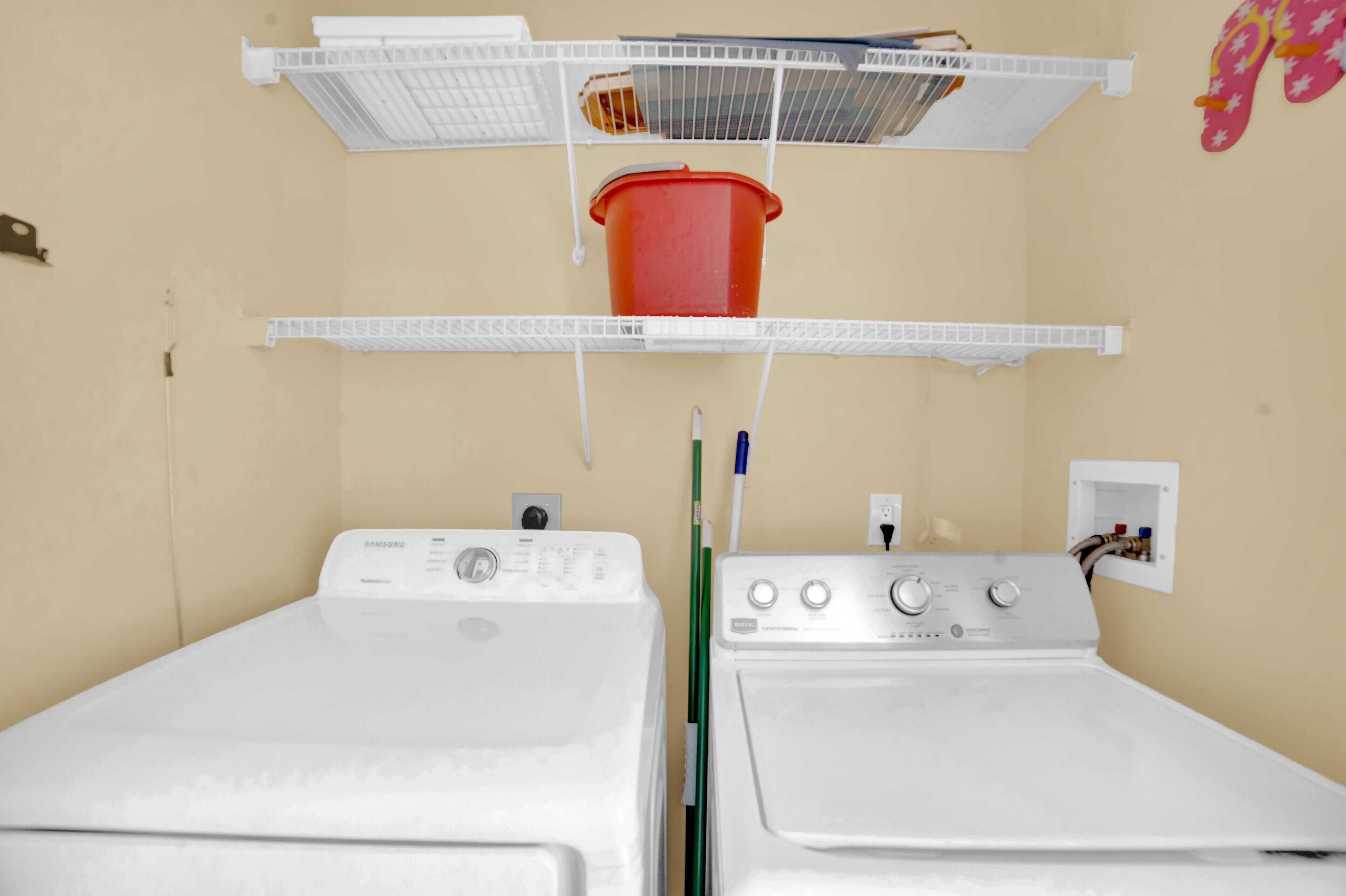 A laundry area with two washing machines, a red bucket, and a shelf with various items.
