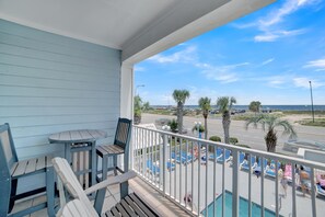 A balcony with a pool, chairs, and a table overlooking a beach and parking area.