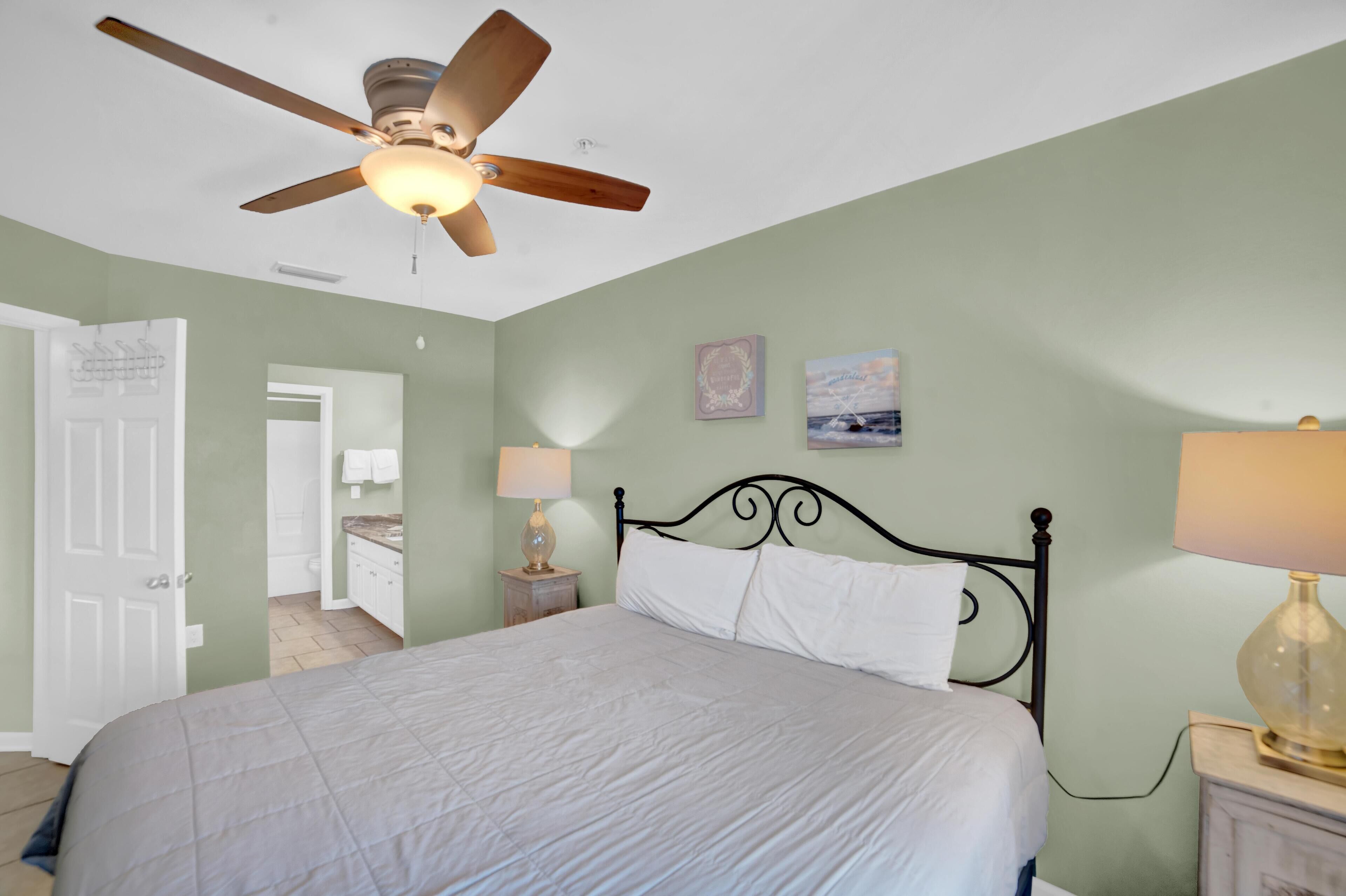 A bedroom with a bed, a ceiling fan, a nightstand with a lamp, and a door leading to another room.