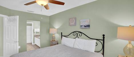 A bedroom with a bed, a ceiling fan, a nightstand with a lamp, and a door leading to another room.