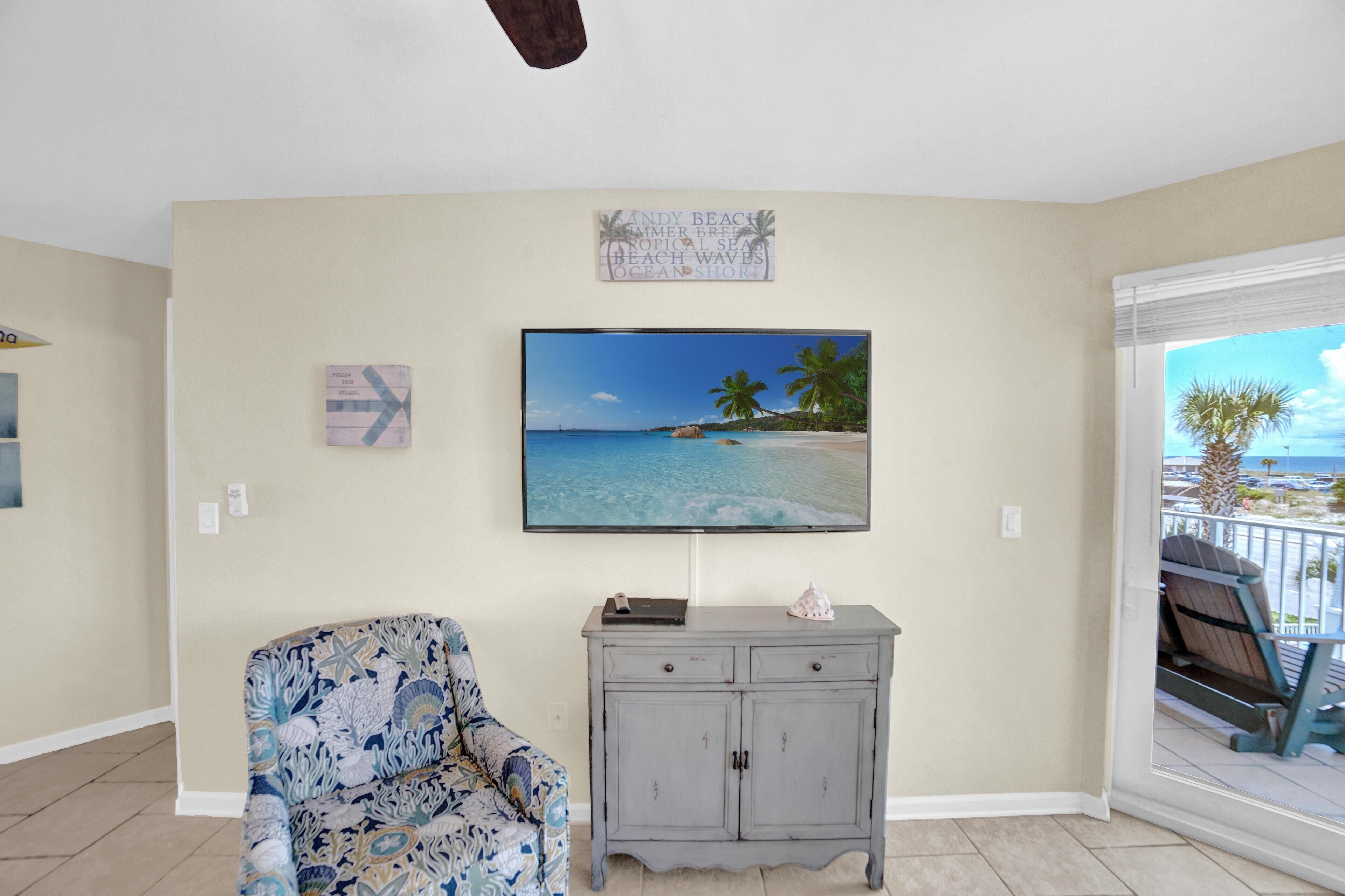 Een woonkamer met een grote televisie waarop een strandscène te zien is, een gestreept fauteuil en een grijze kast.