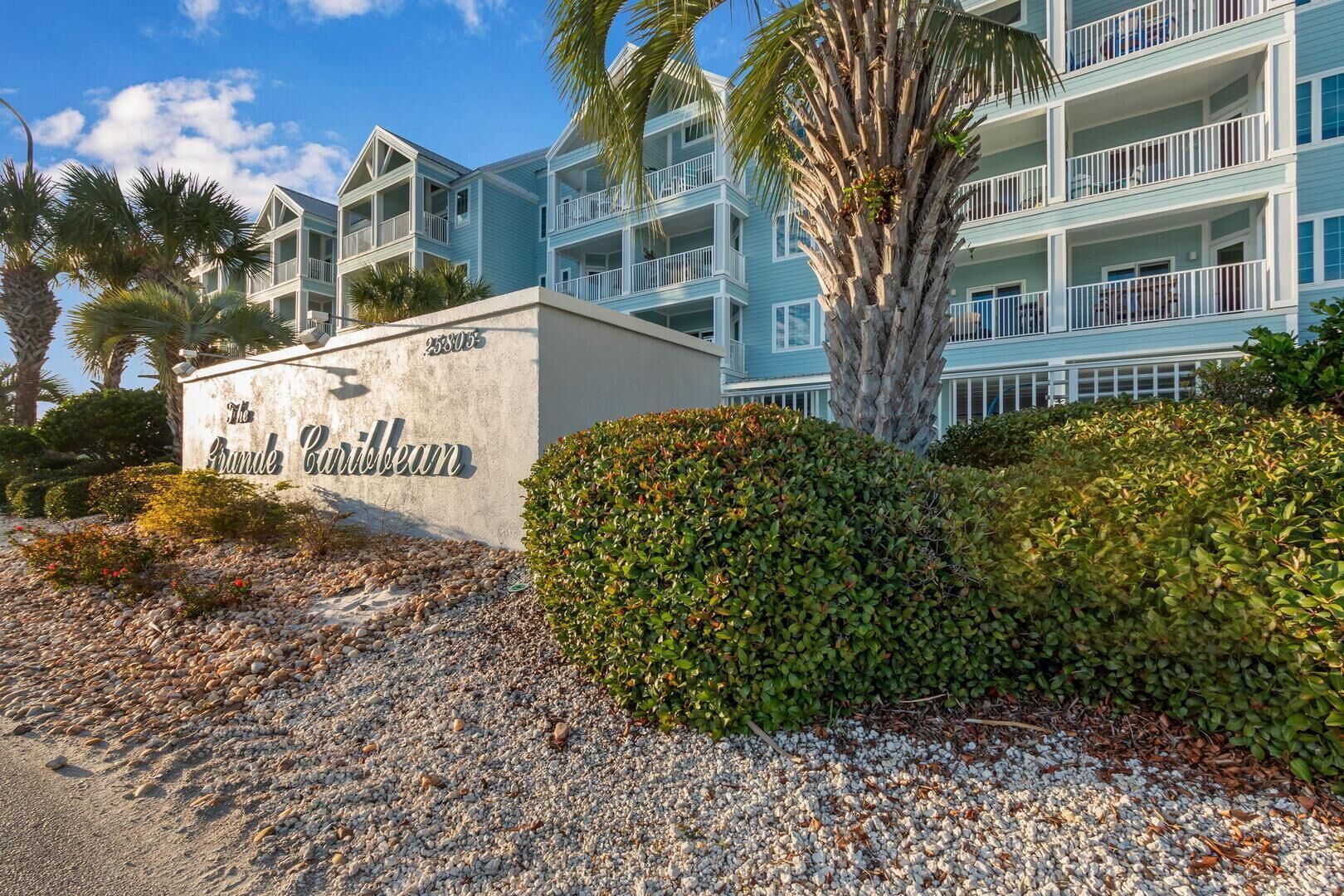 A beachfront hotel with a sign reading "The Brande Caribbean," featuring a multi-story building with balconies and a palm tree in the foreground.