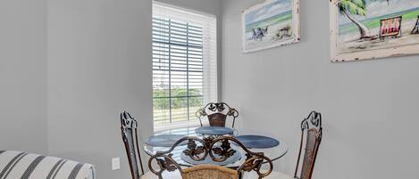 A dining area with a round glass table, wicker chairs, and a window with blinds.
