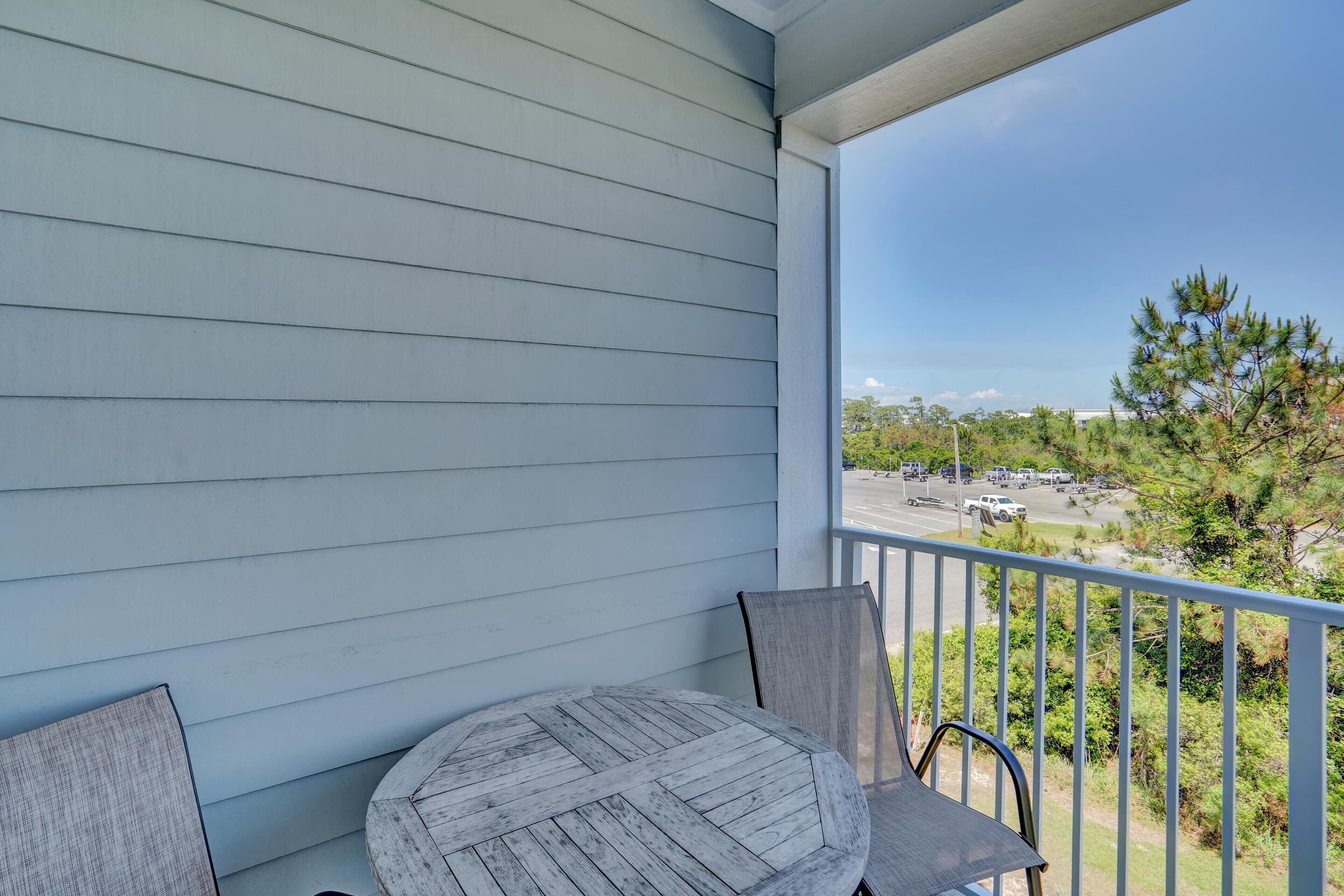 A balcony with a round table and two chairs, overlooking a parking area and trees.