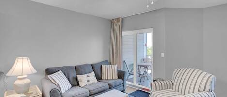 Una sala de estar con un sofá gris, un sillón a rayas y un reposapiés. Hay una lámpara sobre una mesita auxiliar, un ventilador de techo y vistas a un área exterior a través de un ventanal.