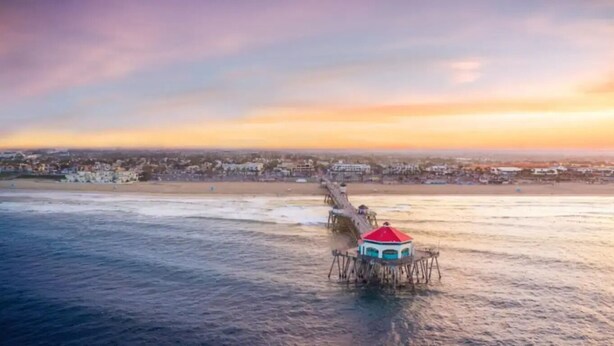 Escena costera con un muelle que se adentra en el agua, una playa y un paisaje urbano al fondo.