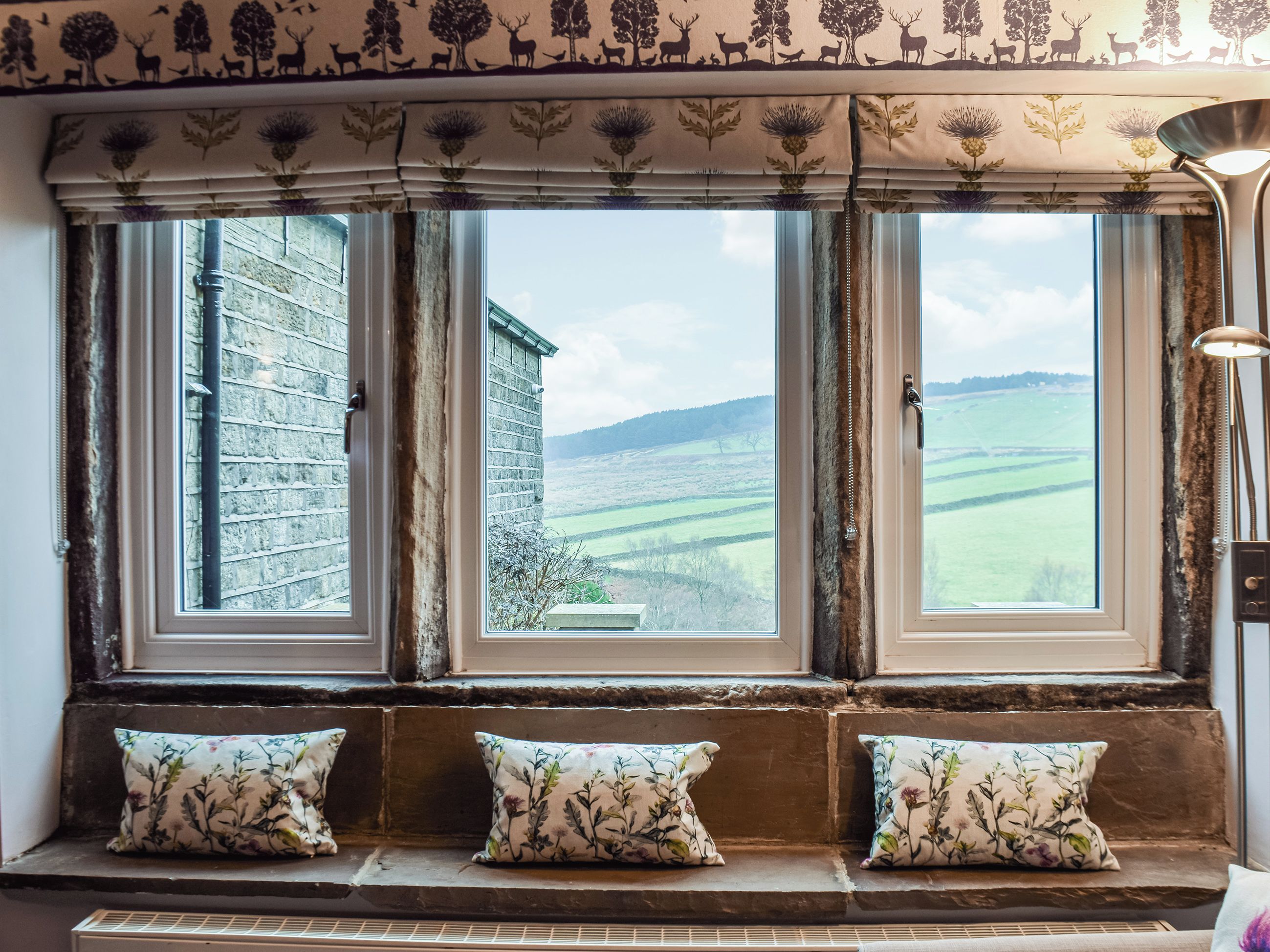 A room with three windows overlooking a rural landscape, a stone bench with cushions, and a lamp on the right.