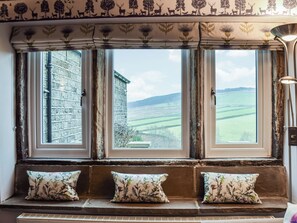 A room with three windows overlooking a rural landscape, a stone bench with cushions, and a lamp on the right.